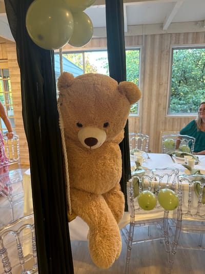 The image depicts an indoor scene, possibly a banquet hall or dining area, decorated for an event. A large, fluffy, light brown teddy bear is prominently featured, suspended by a thick rope and draped with dark green fabric. The bear's arms are hanging loosely, and its body is positioned as if sitting.
In the background, there are several tables set for a meal, with white tablecloths, plates, cups, and green napkins. Transparent chairs surround the tables. Two pale green balloons are tied to the backs of two chairs in the middle ground.
Through large windows in the background, lush green trees and foliage are visible, suggesting an outdoor setting beyond the building. A woman wearing a green top is seated at one of the tables, looking to her left. Another person, partially visible on the far left, is wearing a patterned dress and standing near a table.
The walls of the room are paneled with light-colored wood, and the ceiling has exposed white beams. The lighting appears to be natural, coming from the windows, and possibly supplemented by artificial light. There is no explicit indication of the location in Hem, France within the image itself.