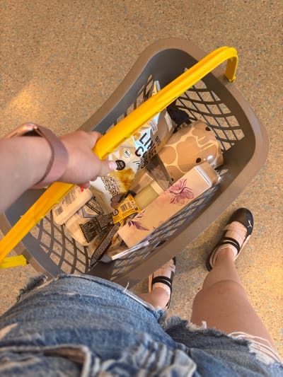 A person's legs and arms are visible in this medium shot, holding onto the yellow handle of a grey shopping basket. The basket contains various items, including a package of "BIO LINSE CHIPS" with German text, a bottle of "WHITE VANCEL & OHNE ALKOHOL," and a box with a floral design. The person is wearing ripped denim shorts and light-colored sneakers with black accents. The scene appears to be indoors, possibly a store, with a speckled floor visible. The lighting suggests it could be daytime. There is no indication of weather or a specific time of day beyond "daytime." The overall impression is of someone shopping. Visible text includes "Natürlich LECKER," "BIO," "LINSE," "WHITE VANCEL," "& OHNE ALKOHOL," and a website address. The location is specified as Erlenbach am Main, Germany.