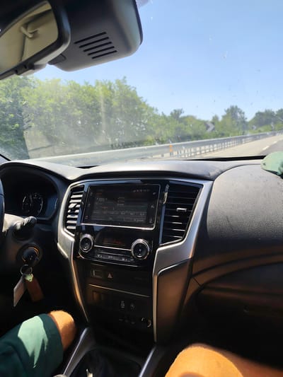 The image shows the interior of a car, likely a pickup truck or SUV, from the perspective of the passenger or rear passenger seat. The dashboard is visible, featuring a central infotainment screen displaying radio information with RDS active, a volume knob, and other buttons. To the left, part of the steering wheel and a gauge cluster are visible. The car appears to be in motion on a road with trees and a clear blue sky visible through the windshield. The windshield has some smudges and what appears to be a small spider web with a spider. A person's leg wearing green shorts is visible in the lower left of the frame, and another person's leg is visible in the lower right, suggesting at least two occupants in the vehicle. A set of keys with a keychain is hanging from the ignition. The environment suggests a daytime scene on a highway or main road. Based on the metadata, the image was taken in Montanaro, Italy.
