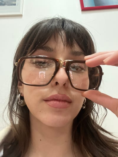 A close-up, chest-up image depicts an individual, likely female, holding eyeglasses in Le Pré-Saint-Gervais, France. The subject has dark brown hair with bangs, light brown eyes, and light pink lips. They wear silver-toned hoop earrings; the right earring includes a small dangling element. The subject holds a pair of large, rectangular, dark brown framed glasses with gold-colored temple accents directly in front of their eyes, gripping the right frame with their right hand (viewer's left). A clear reflection of a person's face is visible in the left lens (viewer's right), and a less distinct reflection in the right lens (viewer's left). The background is a plain, light-colored wall, featuring a partially visible framed picture with a light frame and muted blue/grey content in the upper left, and another with a red frame and blue/turquoise content in the upper right.