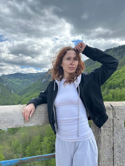 A medium shot shows a woman leaning against a concrete barrier with a landscape of mountains, trees, and sky in the background. The sky is filled with grey clouds. The woman is centered. She has red hair and is wearing a white shirt, grey sweatpants, and a black jacket. Her right arm is bent, with her hand near her head. Her left hand is resting on the barrier. The location is Đurđevića Tara, Montenegro. A blue metal barrier is visible on the right.