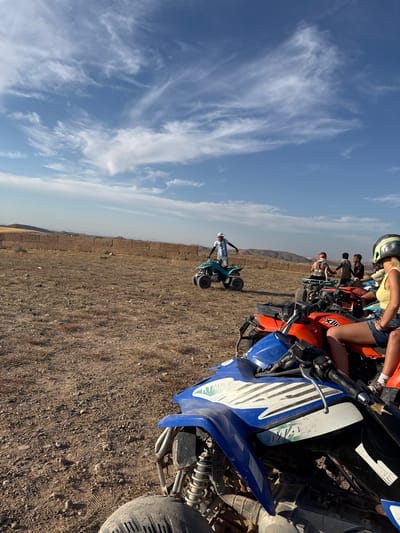 In Lalla Takarkoust, Morocco, a group of individuals are gathered around several all-terrain vehicles (ATVs) in a dry, open field under a bright blue sky with wispy clouds. In the foreground, a blue and black ATV is partially visible, with a person's legs and feet in denim shorts and white socks wearing sneakers seated on an orange ATV behind it. Other ATVs, including an orange one and a teal one, are parked nearby.
A man in a white and blue striped shirt and white cap stands on the teal ATV, gesturing with his arms. Several other individuals, some wearing helmets, are either seated on or standing near the ATVs. In the background, a low, rough-textured wall made of earth or stone runs horizontally across the landscape. Beyond the wall, rolling hills and mountains are visible under the clear sky. The ground is dry and covered with pebbles and sparse vegetation. The overall scene suggests an outdoor recreational activity, likely a tour or adventure involving ATVs in a rural Moroccan setting.