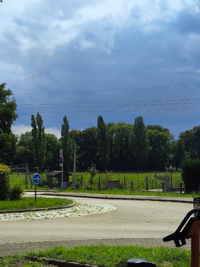 The scene depicts an outdoor environment in Oberschaeffolsheim, France, under a partly cloudy sky with darker clouds dominating the upper left portion. In the foreground, an asphalt road curves to the left around a small, raised circular island paved with light-colored cobblestones arranged in a concentric pattern. Green grass verges line the immediate edges of the road.
On the left side of the road, positioned near the cobblestone island, stands a blue circular road sign with a white arrow pointing right, indicating a mandatory direction. Further along the road on the left, a vertical pole displays two signs: an upper circular sign with a red border and white background showing "30," indicating a 30 km/h speed limit, and a lower circular sign with a red border and blue background, indicating a no-parking zone. Behind these signs, a small, dark utility structure, possibly a shed, is partially visible, along with a public waste bin.
Beyond the road and utility structures, a vast green field extends towards a dense treeline. The field is enclosed by a multi-strand fence. Various wooden or concrete posts are scattered along the field's perimeter. On the right side of the field, a streetlamp with a dark, pyramidal top is visible above a triangular warning sign. Overhead, multiple dark utility lines stretch horizontally across the sky above the treeline. The treeline itself consists of a mix of tall, slender trees, possibly poplars, interspersed with denser deciduous foliage. In the extreme lower right foreground, a portion of a dark-colored outdoor exercise equipment, featuring a black seat, is visible. No people or animals are present in the scene.