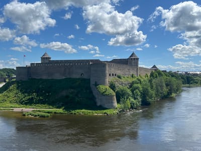 The image shows the Narva Castle, also known as Hermann's Castle, located on the right bank of the Narva River in Narva, Estonia. The castle is a large, stone fortress with multiple towers and high walls, situated on a hill overlooking the river. The sky is blue with white, fluffy clouds. The river in the foreground is wide and dark, with ripples on its surface. Lush green trees and vegetation surround the castle and its embankment. In the distance, across the river, there is a glimpse of the Ivangorod Fortress on the Russian side, and a few buildings of a town. A flag, likely the Estonian flag due to its red, white, and blue colors, is visible on a flagpole near the castle. The scene depicts a historical landmark under a bright, sunny sky.