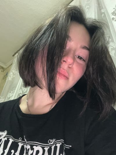 A young person with dark hair and fair skin lies in repose. Strands of hair fall across their face, partially obscuring one eye. The person wears a black t-shirt with stylized white print. A delicate silver chain rests around their neck, and a patterned wallpaper is visible in the background. The image is composed at an angle, giving a sense of informality and relaxation.