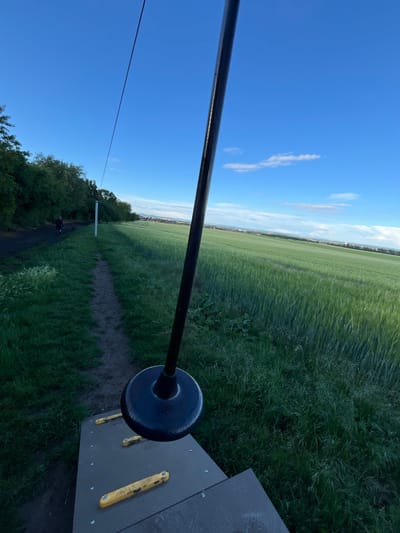 This image captures a view from a zipline starting point in Bašť, Czechia, looking out over a green field under a blue sky with scattered clouds. In the foreground, a black zipline pole with a black circular brake mechanism is visible, attached to a gray platform with yellow handles. A dirt path runs alongside the field, with trees and vegetation bordering it to the left. In the distance, a person is walking a dog on the path, and a town or village is visible on the horizon. The field appears to be planted with a crop, possibly barley or wheat, and it stretches towards the distant settlement. The lighting suggests it is late afternoon or early evening.
