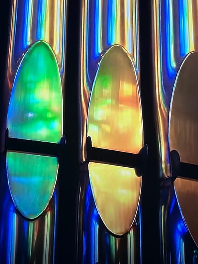 The scene depicts a close-up view of a series of three highly polished, vertical metallic pipes or columns, located in Madzherito, Bulgaria. Each pipe features a large, vertically oriented, elliptical or shield-shaped reflective panel affixed to its upper section. The leftmost visible panel exhibits prominent green and aqua chromatic reflections, while the two panels to its right display strong golden-yellow and orange chromatic reflections. These panels are secured to the pipes by two thin, horizontal, dark-colored support struts each. The vertical pipes themselves show iridescent, multi-colored reflections along their length, primarily featuring streaks of blue, yellow, and orange. The immediate background is uniformly dark. No individuals, actions, or direct interactions are visible in the frame.