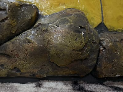 The image shows a close-up, eye-level view of a textured surface, likely artificial rocks, with a bright yellow background. The rocks are dark brown and olive green, with a wet, shiny appearance, suggesting they are either newly made, exposed to water, or covered in a glossy sealant. Some rocks have rough, uneven surfaces with visible pores and crevices. A dark, possibly black, shadow or gap is present between the rocks and the yellow background. The bottom of the image shows a fuzzy, grey and black striped surface, resembling carpet or fabric. No people or identifiable landmarks are visible. The provided location of Kalininskaya, Russia, does not appear to be reflected in the visual information of the image.