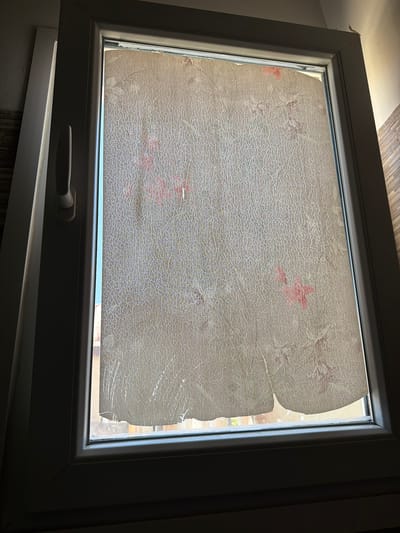 The image shows a close-up view of a white window frame with a slightly aged and cracked translucent film applied to the glass. The film has a faint pattern of pinkish floral designs and a distinct crackled texture. The bottom edge of the film is torn and peeling away from the glass, revealing the outside environment. The window is situated indoors, with interior wall textures visible to the left and right. The lighting suggests it is daytime, with light filtering in from outside. The scene is likely a residential or commercial interior in Dromolaxia - Meneou Municipality, Cyprus, and the condition of the film suggests a need for repair or replacement. There are no people visible in the image.