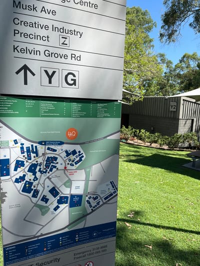 The image captures a vibrant outdoor scene on a sunny day at the Queensland University of Technology (QUT) Kelvin Grove campus in Australia. The primary subject is a large, grey information sign and campus map, positioned upright, with a bright blue sky and lush green trees visible in the background.
The left panel of the sign provides directional information and listings. It points with an arrow to "Y G" and lists "Musk Ave," "Creative Industry Precinct Z," and "Kelvin Grove Rd." Further down, sections detail "FACULTIES" (Creative Industries Faculty, Faculty of Education) and "INSTITUTES," alongside "BUILDING" codes (21-26, 29-213, E, A, B, K, N, U) for various services such as Accommodation, Equity, Student Counselling, Food Court, and HIQ.
The right panel of the sign displays a detailed campus map of QUT Kelvin Grove, highlighting "YOU ARE HERE" with a red dot. The map outlines campus buildings, roads like "INNER NORTHERN BUSWAY," and surrounding areas such as "Victoria Park Golf Course," "QUT Kelvin Grove Busway Station," "Village Centre," "U 44 Musk Avenue," and "McCeskie Park." A legend explains symbols for parking, toilets, child care, water, AED, bike parking, bus stops, and taxis. Below the map, emergency contact numbers for "Emergency 3138 8888" and "Freecall 1800 065 585" are displayed, along with "QUT Security." A list of buildings with letter codes (e.g., A, B, C) includes locations like QUT Art Museum, QUT Bookshop, QUT Medical Centre & Pharmacy, HIQ, The Grove Bar, and Sports Court.
Behind the sign, a well-manicured patch of green grass is visible, with distinct shadows suggesting bright sunlight. Beyond the grass, a modern building with horizontal siding and a "Y2" sign on its wall can be seen. No people are present in the frame, emphasizing the architectural and natural elements of the campus environment. The clear blue sky and bright illumination indicate pleasant weather conditions.