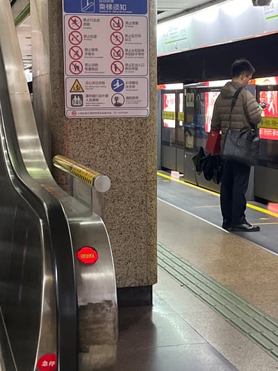 The scene depicts a subway station platform in Yanqiao, China. In the foreground, the silver-metallic side of an escalator is visible, featuring a red circular light displaying a horizontal dashed pattern, likely indicating "no entry" or "stopped," with a red "急停" (Emergency Stop) button below. A textured granite-like pillar stands center-left, bearing a white sign detailing "自动扶梯 乘坐须知" (Customs for Taking the Escalator) with pictograms in Chinese and English. To the right, a male individual stands on the platform facing closed subway train doors. He wears a light brown puffer jacket, dark trousers, and dark shoes. He carries a dark patterned shoulder bag on his left side, and in his left hand, he holds a red rectangular object and several dark bundled items, while observing a luminous handheld device. The platform edge is marked by a yellow safety line, and tactile paving is integrated into the tiled floor. The train is stationary, its platform screen doors displaying interior lighting and visual information.