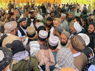 The image shows a crowd of men gathered in a tent in Zaker-E Sharif, Afghanistan. The men are mostly wearing traditional Afghan clothing, including turbans and long robes. Some of the men are talking to each other, while others are looking at the camera. There is a man in the center of the image holding a camera up to his face to take a picture. The image was captured at the coordinates 31.547 latitude, 65.761 longitude, which is located in Zaker-E Sharif, Afghanistan.