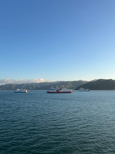 The image depicts a serene seascape with three ferries navigating the waters. The ferries are positioned at varying distances from the viewer, with the closest one being a red and white vessel, followed by a white ferry with blue accents, and a third white ferry further in the distance. The water is calm with gentle ripples. In the background, lush green hills and mountains rise, partially covered by wispy clouds. The sky is clear and a vibrant blue, suggesting a pleasant day. The location is identified as Batas Desa Skotong Timur, Indonesia. There are no people visible in the image.