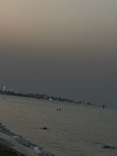 The image depicts a twilight scene at a beach in Sousse, Tunisia. The sky is overcast and a muted gray, suggesting either dusk or dawn. In the distance, a shoreline is visible with a scattering of buildings and lights, indicating an urban or developed coastal area. Several illuminated structures, possibly decorative lights or towers, stand out along the coastline. The sea is calm with gentle ripples and waves lapping at the sandy shore in the foreground. Several figures are present in the water; two individuals appear to be swimming or wading near the middle ground, and another dark shape, possibly an animal or person partially submerged, is visible closer to the shore. A black buoy or marker stands upright in the water further out. The overall atmosphere is serene and quiet, typical of a beach at the edge of the day.