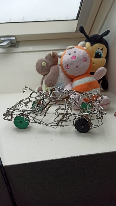 The image captures an indoor scene, likely a collection of items displayed on a surface next to a window. The image appears to be rotated 90 degrees counter-clockwise from its natural orientation.
On the left side of the frame, a window with a white frame and a metallic latch is visible. The view outside the window is bright and white, suggesting an overcast sky or bright daylight.
In the center and top-left, several plush toys are stacked or placed closely together. The topmost plush toy is a black and yellow bee-like creature with prominent blue eyes and long eyelashes, and what appear to be antennae. Below it is an orange and white plush toy featuring a round face with pink cheeks and spiral patterns on its head. A brown plush toy is partially visible beneath the orange one, with a light green circular detail on one of its limbs.
To the right of the plush toys, a complex structure made of metal wire dominates the right half of the image. This intricate wire structure appears to be a sculpture or a decorative object, possibly resembling a vehicle or an abstract form. It features small wheels; one distinct green textured wheel is visible towards the front/bottom of the structure, and another black textured wheel is visible towards the top/back.
The items are resting on a light-colored, possibly white or off-white, flat surface, which could be a shelf or a tabletop. In the bottom right corner, a dark object or shadow is visible, forming a diagonal line. Faint white text is discernable on this dark surface, which appears to read "KEMA-KEUR" followed by other characters, often a certification mark found on electrical products or cables.
The overall scene is a still life, showcasing personal items in a casual arrangement, illuminated by natural light coming from the window. No specific activity is taking place.