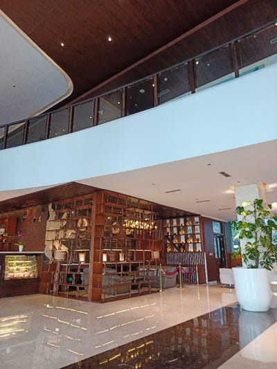 A well-lit hotel lobby features a high ceiling with a dark wood design. A second-floor balcony with glass railings overlooks the main floor. A large wooden shelving unit filled with decorative items separates the seating area. A dessert display sits on the left. A potted plant rests on the right. The polished floor reflects the light and elements of the lobby.