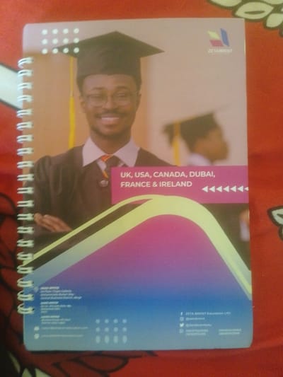 The image is a close-up, overhead shot of a spiral-bound notebook. The notebook's cover features a graduation-themed design with a young Black man wearing a graduation cap and gown, smiling at the camera. The background of the cover is a gradient of pink, purple, and blue. Text on the cover reads "UK, USA, CANADA, DUBAI, FRANCE & IRELAND" and lists contact information for "ZETA BRENT Education LTD". The notebook is lying on a textured red surface. The location in the city of Magajin Gari, Nigeria, is not depicted in this image.