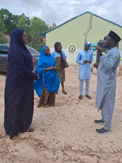 A group of people are gathered outside a building in Kundulun, Nigeria. Two men, dressed in traditional Nigerian attire, stand in the foreground. The man on the left wears a light grey thobe and a black cap, while the man on the right wears a light blue thobe and a patterned cap. They appear to be conversing, with the man on the left gesturing with his hands.
Behind them, three women are also present. The woman closest to the foreground wears a dark purple hijab and dress. Next to her, a woman in a bright blue hijab and patterned skirt is visible. Another woman, wearing a black hijab and a blue and patterned top, stands behind her.
The setting is a sandy area in front of a light green building with a dark green roofline. A crack runs down the face of the building. There is some greenery and trees in the background. A car is partially visible on the left side of the image. The sky is overcast, suggesting it might be a cloudy day. The time of day appears to be daytime. No specific event is evident, and the mood seems neutral. A small circular emblem with a green and gold design is visible on the building. There is some text on the building that appears to say "SAL STATE" which likely refers to the state in Nigeria.