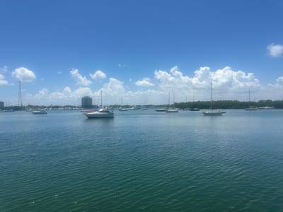 The image depicts a wide view of the bay in North Bay Village, Florida, under a bright blue sky dotted with fluffy white clouds. Several boats of various sizes are moored on the calm, blue water, creating a serene marina scene. In the background, a dense line of green trees separates the water from the city skyline, which features a tall, slender tower and a few buildings. The water itself is slightly rippled, reflecting the sunlight and adding texture to the scene. There are no people visible in the image.
