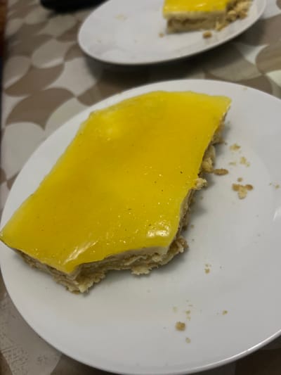 This scene is situated in Ksar-el-Kebir, Morocco. The image displays two partially consumed dessert slices on white circular plates, positioned on a geometrically patterned table surface. In the foreground, a rectangular slice of dessert, approximately 10x6 cm, is centered on a white plate. This dessert features a bright yellow, translucent, slightly textured top layer, suggestive of lemon curd or glaze. Beneath this, a layered or crumbly light-beige base is visible, consistent with a shortbread or biscuit crust. A noticeable bite has been taken from the left side of the dessert, exposing the internal structure. Several small, light-colored crumbs are scattered on the white plate surface surrounding the main dessert. In the blurred background, a second white plate holds another similar, partially eaten dessert slice. The table underneath exhibits a repeating pattern of light brown and off-white interlocking shapes. No individuals are visible.