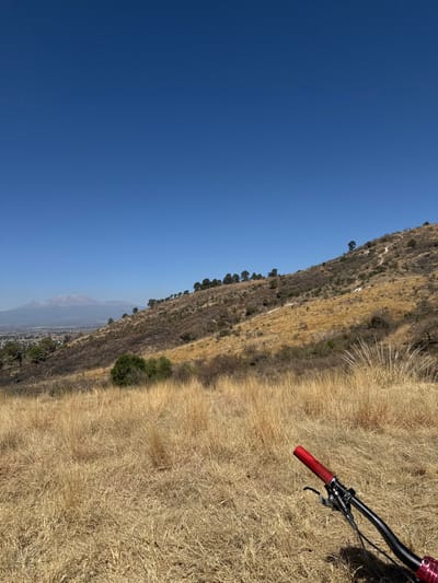 The scene depicts a sunlit, arid hillside overlooking the city of San Gregorio Zacapechpan, Mexico, and distant snow-capped mountains under a clear blue sky. In the immediate foreground, positioned in the lower right, is the black handlebar of a bicycle, featuring a prominent red grip on the right side, along with visible brake levers and cables. The ground in the foreground and midground consists of dense, dry, golden-brown grasses and scattered low-lying shrubs. A steep hillside, covered in similar dry vegetation interspersed with small dark green trees, ascends towards the upper right. A narrow, winding dirt trail is visible traversing this hillside. In the background, beyond the hillside, a sprawling urban area identifies as San Gregorio Zacapechpan, partially obscured by atmospheric haze. Farther in the distance, a large mountain range with snow-capped peaks is visible against the horizon. No individuals are explicitly present, but the inclusion of the bicycle handlebar suggests a user's perspective during a stop. The environment indicates dry, mountainous terrain under bright, clear conditions.