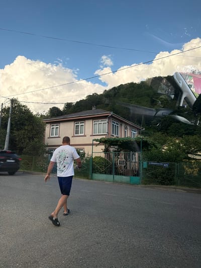 In a roadside scene in Krayevsko-Armyanskoye, Russia, a person is captured mid-stride, walking on an asphalt road. The individual is wearing a white t-shirt with a graphic print and dark blue shorts, along with black slip-on sandals. Their posture suggests a casual walk or perhaps a playful pose, with one arm extended backward and the other forward.

The background features a two-story building with a light-colored facade and white-framed windows, set against a backdrop of lush green trees that climb a hillside. A green metal fence runs along the property line, with a gate visible. A blue street sign displaying "65" and Cyrillic text, "ул. Измайловская," indicates the location.

The sky above is a clear, bright blue, interspersed with fluffy white clouds, suggesting a sunny day, likely in the late morning or early afternoon. Power lines stretch across the frame, a common sight in many residential areas. The overall impression is a snapshot of everyday life on a pleasant day in a semi-urban or suburban environment. The reflection in what appears to be a car windshield, which is visible in the upper part of the image, shows the sky and the surrounding buildings.