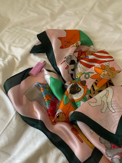 The scene is set indoors, within a private residence or lodging, stated to be in Πλατύς Γιαλός, Greece. A colorful, intricately patterned silk scarf is loosely draped across a wrinkled white fabric surface, consistent with a bed sheet or duvet cover. The scarf features a light pink background with a dark green border. Its pattern depicts various anthropomorphic and cartoonish cats in different poses and costumes, including an orange cat with a green lower body, a cat in a black and white cow-print costume, a cat with orange and white stripes, a cat with an orange face and a yellow sun-like collar, a cat in an orange leafy costume, and a cat head emerging from a shark costume. A small, pink, rectangular object, identified as a "Clipper" brand lighter, rests horizontally on the scarf near the upper left quadrant. No individuals are present, and no actions or interactions are depicted. The lighting is diffuse and ambient.