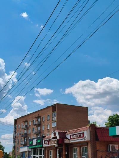 This image captures a street view in Velyka Bahachka, Ukraine, on a sunny day. A multi-story brick apartment building dominates the background, with several floors featuring small balconies. Below the apartment building, a row of storefronts is visible, including one with a sign that reads "ПРОДУКТИ," indicating a food store. Power lines stretch diagonally across the bright blue sky, which is dotted with fluffy white clouds. The scene suggests a typical urban environment in a Ukrainian town, with a mix of residential and commercial structures.