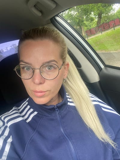 A woman with blonde hair wearing round-frame glasses and a blue Adidas-striped jacket is sitting in a car. Raindrops are visible on the car window, which is behind her. Outside the window, trees and a red fence are visible. This scene is likely taking place in Danilov, Russia.
