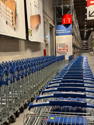 This image depicts the interior of an IKEA store's self-service furniture pickup area in Corsico, Italy. The scene is dominated by multiple long, parallel rows of blue-handled metal shopping carts, designed for transporting flat-pack furniture, extending from the foreground into the midground. To the left, a light-colored brick wall features two large white banners displaying IKEA products: one illustrating stacked brown boxes forming a drawer unit, and another showing wall-mounted wooden hooks. An IKEA logo is visible on the lower section of the second banner.

Overhead, a red cube sign labeled "Scaffale 1 1" (Shelf 1 1) is suspended from the ceiling structure. Below it, a blue sign reads "BENVENUTI al Self Service mobili." (WELCOME to the mobile Self Service.), with a white instructional sign beneath it detailing shelf locations ("Lo scaffale 00 lo trovi lungo il percorso e nell'area prima delle casse" - Shelf 00 can be found along the path and in the area before the checkouts) and a diagram illustrating a layout of shelves (Scaffale 00 Posto 01-04).

In the background and extending to the right, tall industrial shelving units filled with various boxed products are visible under bright overhead lighting. No individuals or active interactions are present in the image. A red fire extinguisher box is mounted on the left wall. The floor is light-colored concrete.