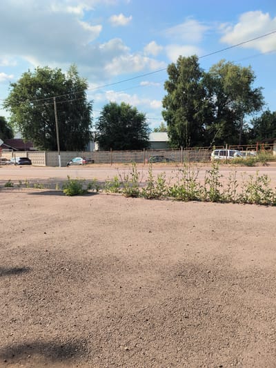 The image depicts a roadside scene in Pokhvistnevo, Russia, under a partially cloudy blue sky. In the foreground, a gravelly ground surface dominates the lower portion of the frame, with sparse weeds and vegetation growing along a raised edge. Beyond this is a paved road with a white line marking the lane.

Across the road, a concrete fence runs horizontally, with a metal fence with orange bars to its right. Behind the fences, a row of trees forms a dense green backdrop. Several vehicles are visible: a dark-colored sedan parked on the left behind the concrete fence, a silver sedan slightly further right, and a white van on the far right. A single utility pole stands tall amongst the trees on the left side. The overall impression is one of a suburban or semi-rural roadside on a clear day.