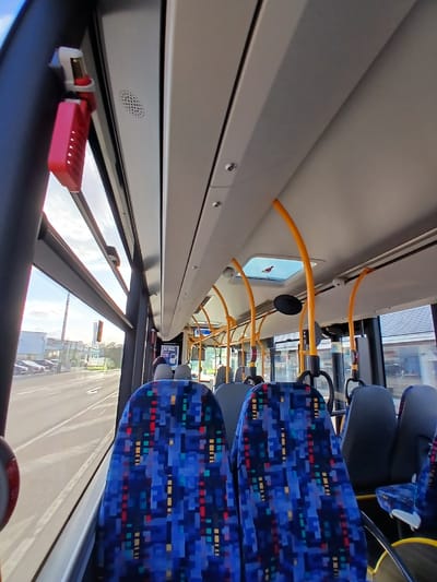 The scene depicts the interior of a public transport bus, observed from a mid-section viewpoint looking forward, while traversing an urban or industrial area in Homburg, Germany.

The bus interior features light gray overhead panels with integrated lighting and a central rectangular skylight. Multiple curved, yellow grab poles extend from the ceiling, equipped with black horizontal handholds. On the left side, affixed to a window support pillar, is a red emergency hammer secured in a white plastic holder, positioned above a circular, perforated speaker grill. Rows of passenger seats are visible, primarily upholstered in a distinctive blue fabric patterned with pixelated blocks of red, yellow, and teal. The seatbacks are gray. In the mid-distance, a circular convex mirror for the driver is mounted overhead, and an illuminated display screen is visible on the right side of the aisle. The bus appears to have sparse occupancy, with faint outlines of individuals visible seated towards the front.

Through the large windows on the left, an exterior environment is visible under a light blue sky with scattered white clouds. An asphalt road with white forward-pointing arrow lane markings runs parallel to the bus. Along the roadside, dark-colored cars are parked in front of industrial-style buildings, including one prominent white structure. A tall, slender, reddish-brown industrial chimney or stack is visible in the distance, alongside a partially obscured blue vertical sign displaying the word "Autohaus."
