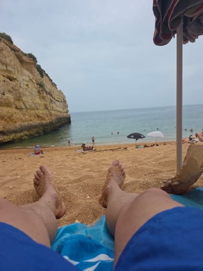 The image depicts a first-person perspective of someone relaxing on a sandy beach in Porches, Portugal, likely during daytime. The foreground is dominated by the legs and feet of the individual, covered in sand, resting on a blue towel. To the right, part of a striped beach umbrella is visible, along with a crumpled paper bag. The middle ground shows a sandy beach with several people sunbathing and walking. Two beach umbrellas, one black and one white, are set up in the sand. The sea is calm with several individuals swimming or wading in the water. In the background, a large, light-brown cliff face with some vegetation on top juts out into the sea on the left side of the frame. The sky is overcast with a light-grey hue.