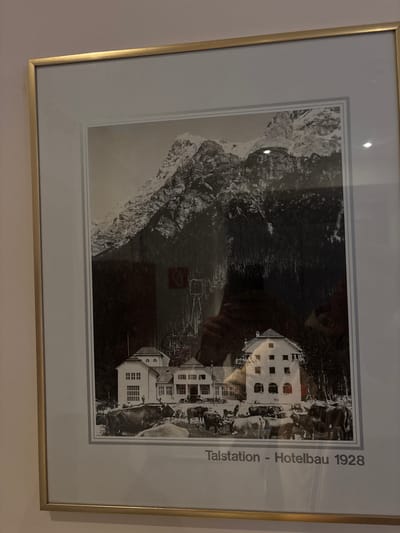 A framed black and white photograph depicts a scenic mountain village with a prominent hotel. In the foreground, several cows graze peacefully in a field. Behind them, a large, multi-story hotel with a distinctive architectural style stands against a backdrop of lush green trees and a majestic, snow-capped mountain range. The scene is labeled "Talstation - Hotelbau 1928," indicating it is a view of the hotel construction or its base station in 1928. The location is identified as Ehrwald, Austria.