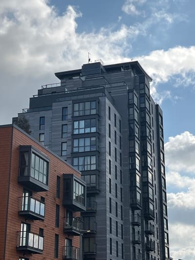 This is a daytime photograph capturing a view of two modern buildings against a partially cloudy blue sky in Slough, United Kingdom. The image, originally rotated, is oriented to show the structures extending upwards.
Dominating the upper and central parts of the frame is a tall, multi-story building featuring a dark grey facade, possibly made of wood paneling or a similar composite material. Its design is contemporary, with numerous windows, some recessed, others protruding like dormers or integrated into sloped roof sections across various levels. The building exhibits a stepped or terraced silhouette, creating distinct horizontal lines and angles. Small details like an antenna or sensor are visible on its upper edge, and a tiny green tree grows on one of its lower terraces.
In the foreground, towards the bottom right, is a shorter building with a reddish-brown facade, likely brick or terracotta, and a sloped roof. This roof is notable for several large, square-shaped skylights or dormer windows that rise prominently from its surface.
The sky overhead is bright blue with scattered white cumulus clouds, indicating fair weather. A small, dark, unidentifiable object, possibly a bird or aircraft, is faintly visible in the upper left portion of the sky near the dark building. No people or specific activities are depicted, and there is no visible text in the image.