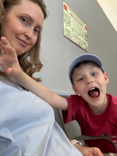 A woman and a young child are posing for a selfie. The woman is lying down, looking at the camera with a slight smile. The child is positioned above her, also looking at the camera with his mouth wide open. He is wearing a red shirt and a blue cap.
Behind them is a wall with a fire evacuation plan printed on it. The plan includes the text "ПЛАН ЭВАКУАЦИИ ПРИ ПОЖАРЕ" and "ЕДИНАЯ СЛУЖБА СПАСЕНИЯ," indicating that the image was taken in Russia. The text on the evacuation plan also includes "Железнодорожный вокзал станция лесовинь," which can be translated to "Railway station station forest," and "112" indicating it is a Russian railway station. The station is most likely in the city of Balezino, Russia, given the other text on the sign.