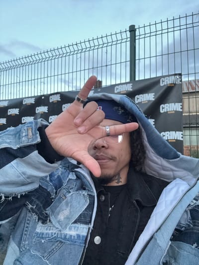 An individual is depicted in a close-up, self-portrait style photograph, situated outdoors in Noisy-le-Sec, France. The subject is a person with dark, curly hair, a dark goatee, and wearing clear-rimmed eyeglasses. They are wearing a blue bandana, a black t-shirt, a black denim jacket, and a distressed, patchwork denim hooded jacket with a white quilted lining. A black tattoo is visible on the left side of their neck. Their right hand is raised, with the thumb extended and index finger pointing upwards, forming an 'L' shape, partially obscuring their face below the eyes. Rings are present on their right index and middle fingers. In the background, a light gray metal panel fence with dark green posts is visible, running horizontally. Below the top of the fence, a black banner is strung, displaying repetitive white text that reads "CRIME" with "SESSION" underneath in smaller font. The sky above the fence is overcast and light blue. Portions of a building with reddish-brown brickwork are faintly discernible behind the banner and fence.