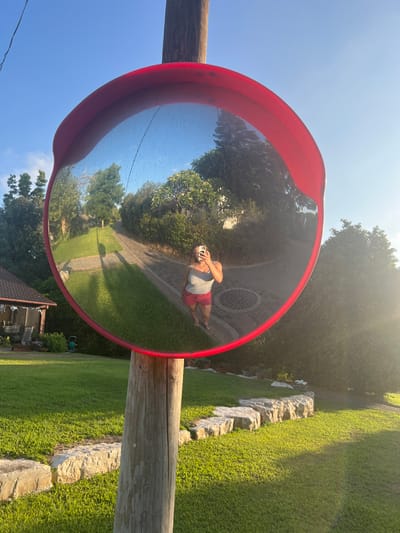 A convex traffic mirror is affixed to a wooden post, reflecting a sunlit scene. The reflection includes a woman taking a photo with a phone, a paved path, and lush greenery. The foreground displays grass and a low stone wall. The background contains trees and a blue sky. The mirror has a red frame.