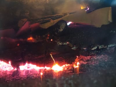 The image displays the interior of a combustion chamber, likely a fireplace or stove, located in Aumetz, France. The upper portion of the frame features several pieces of corrugated cardboard, partially consumed, showing both their light brown, layered structure and areas of black charring with some visible orange and yellow flames. Beneath this, a large, dark, irregular mass of charred material or unburnt fuel forms a horizontal band across the middle. The lower section is dominated by a bed of actively glowing red and orange embers and ash, indicating intense heat and smoldering. Numerous bright orange-yellow sparks are distributed throughout the lower and middle areas, with a distinct upward-streaking spark visible in the lower right quadrant. The scene is illuminated solely by the combustion, creating strong contrasts and a warm color palette against the darker, charred elements.
