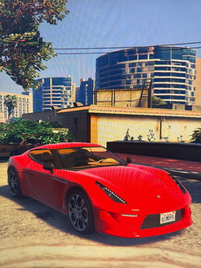 A bright red sports car is parked on a street in a city. The car has a black interior and is facing forward. Behind the car, there are several tall buildings, some with reflective windows. Trees and bushes are visible on the sides of the street, and power lines stretch across the sky. The ground is paved with asphalt. The license plate on the car reads "02 MU 995". The scene appears to be a digital rendering, likely from a video game, due to the pixelated quality of the image. There is no indication of the location being Pavolche, Bulgaria.
