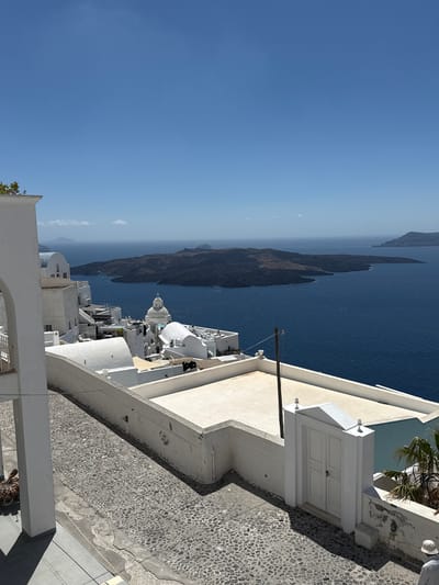 The image depicts a view of the caldera in Santorini, Greece, specifically from the town of Fira. In the foreground, a cobblestone path leads past whitewashed buildings and a white double door. Beyond the buildings, the deep blue waters of the Aegean Sea stretch out, with the volcanic islands of Nea Kameni and Palea Kameni visible in the distance. The sky is clear and bright blue. A few small boats can be seen on the water. The architecture is characteristic of the Cycladic islands, with cubic white buildings, domed roofs, and arched doorways. The overall scene conveys a sense of tranquility and the iconic beauty of Santorini.