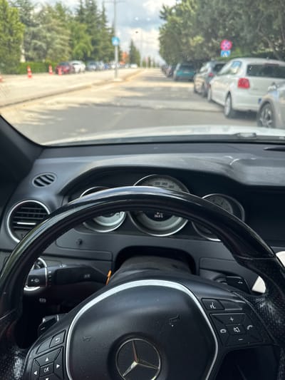 The image is a point-of-view shot from the driver's seat of a Mercedes-Benz. The steering wheel is prominently featured in the foreground. The dashboard displays multiple gauges, including a speedometer and a temperature gauge reading 26.8 degrees Celsius. The view through the windshield shows a street lined with trees and parked cars. Several orange traffic cones are visible on the side of the road. A blue and white pedestrian crossing sign is present. Further down the street, traffic signs including a red circle with a diagonal bar and a red "X" symbol are visible. The overall impression is a typical urban street scene in Etimesgut, Turkey, on a moderately cloudy day.