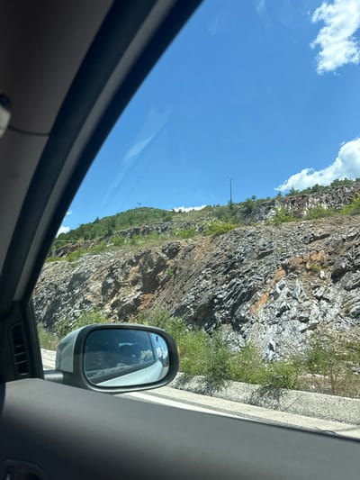 The image captures a view from inside a vehicle, looking out towards a rocky, sloped hillside under a bright blue sky with scattered white clouds. The rock face is a mix of gray, white, and reddish-brown, with vegetation growing sparsely on its surface and at its base. In the foreground, the interior of the car is visible, including the side mirror reflecting the sky and the interior of the vehicle. The side mirror is positioned in the lower left quadrant of the image. The overall scene suggests a drive through a mountainous or hilly area. The location is identified as Kato Nevrokopi, Greece.
