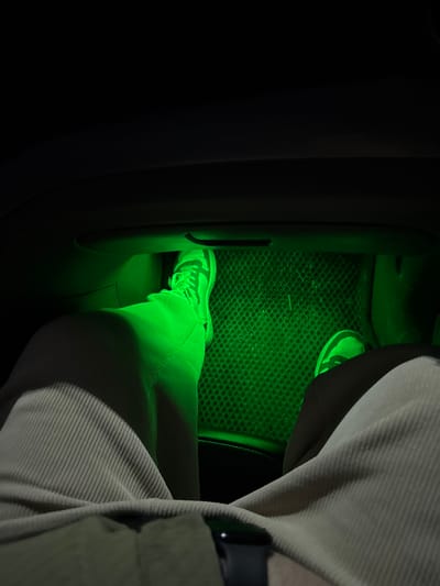 Inside a vehicle, a person's leg and foot are visible. The foot is wearing a sneaker and is placed near a vehicle floor mat with a grid pattern. A dark console is adjacent. The individual is wearing corduroy pants. The scene is bathed in green light, possibly due to interior lighting. A seat is also visible.