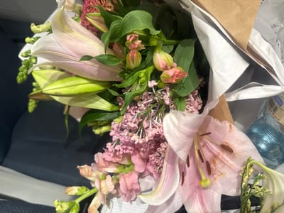 A bouquet of pink lilies and pink stock flowers is displayed in brown and white paper wrapping. The flowers sit on a blue seat, likely inside an airplane. A bottle of water is visible near the flowers. The location is unknown.