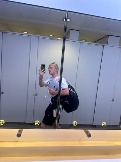 A reflection of a young man with fair hair in a mirror is seen. The man is holding a smartphone in his right hand and a laptop in his left, which is adorned with stickers. He is wearing a white t-shirt, dark track pants, and carrying a bag. The mirror is framed with a metal border and features circular lights with hand symbols. The background appears to be an indoor space with white walls and a series of panels. The image was not taken in Uttlesford, United Kingdom.