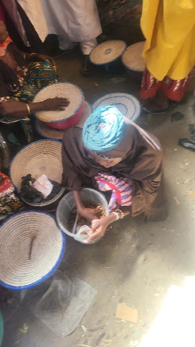 The scene depicts an informal vendor stall or market area in Fagge D1, Nigeria. A central female figure, viewed from an elevated perspective, crouches on a rough, dusty concrete or dirt floor. She wears a brown garment and a light blue head covering. Her hands are submerged in a clear plastic bucket containing small, dark, granular material, and she holds a small clear plastic bag, indicating an action of scooping or packaging. Light-colored bracelets are visible on her wrists. Surrounding her on the ground are multiple large, circular, woven baskets, primarily white with blue rims, some containing dark goods and small white plastic bags. Other individuals are partially visible: a person wearing a patterned garment with a gold bracelet is to the upper left; a person in white attire stands to the upper center-left; and a person wearing a yellow garment over red patterned trousers is to the upper right. The environment appears to be an active, open-air commercial space with strong overhead lighting casting subtle shadows.
