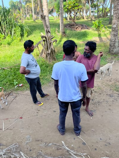 In Moratuwa, Sri Lanka, a group of three men and two dogs are gathered on a dirt path, likely a rural road or clearing. The environment is tropical, with lush green grass, palm trees, and other dense vegetation surrounding the area. The sky is bright, suggesting it is daytime, and the sun casts shadows.

The man on the left, wearing a light grey t-shirt and dark pants with yellow flip-flops, appears to be speaking with the other two men. He has his right hand on his hip. In the center of the image, a man in a white t-shirt and dark pants faces away from the camera, partially obscuring the man behind him. The man on the right, wearing a maroon t-shirt and light-colored shorts with dark sandals, is actively engaged in conversation, gesturing with his left arm. He has a beard and appears to be speaking intently.

Two dogs are present. A small tan and white dog is on the left side of the frame, facing the man in the grey t-shirt. A larger, light-colored dog stands on the right, looking towards the man in the maroon t-shirt. Debris, including leaves and a discarded red and white packet, are scattered on the ground. The overall scene depicts a casual, everyday interaction in a tropical setting.