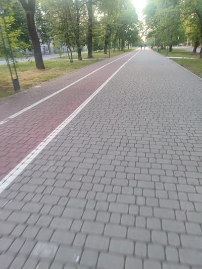 The image shows a paved pathway with a red-colored lane for cyclists or pedestrians, separated by white lines, alongside a gray paved path. Trees line the pathway on the left, and a street with buildings and cars is visible in the background. In the distance, several people are walking or sitting on benches. The scene appears to be a public park or promenade within the city of Zhovti Vody, Ukraine, given the context.
