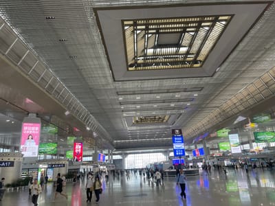 The image depicts a spacious indoor area, likely a transportation hub like an airport or train station, in Yuhuatai District, China. A large number of people are seen walking through the polished floor, some wearing masks. The ceiling is high and features a complex design of gridded metal panels, interrupted by a square, multi-layered opening revealing the structure above. Signage in Chinese and English hangs from the ceiling, providing information about gates and advertisements for various products and services. The light is bright and uniform, reflecting off the floor and creating a sense of openness.
